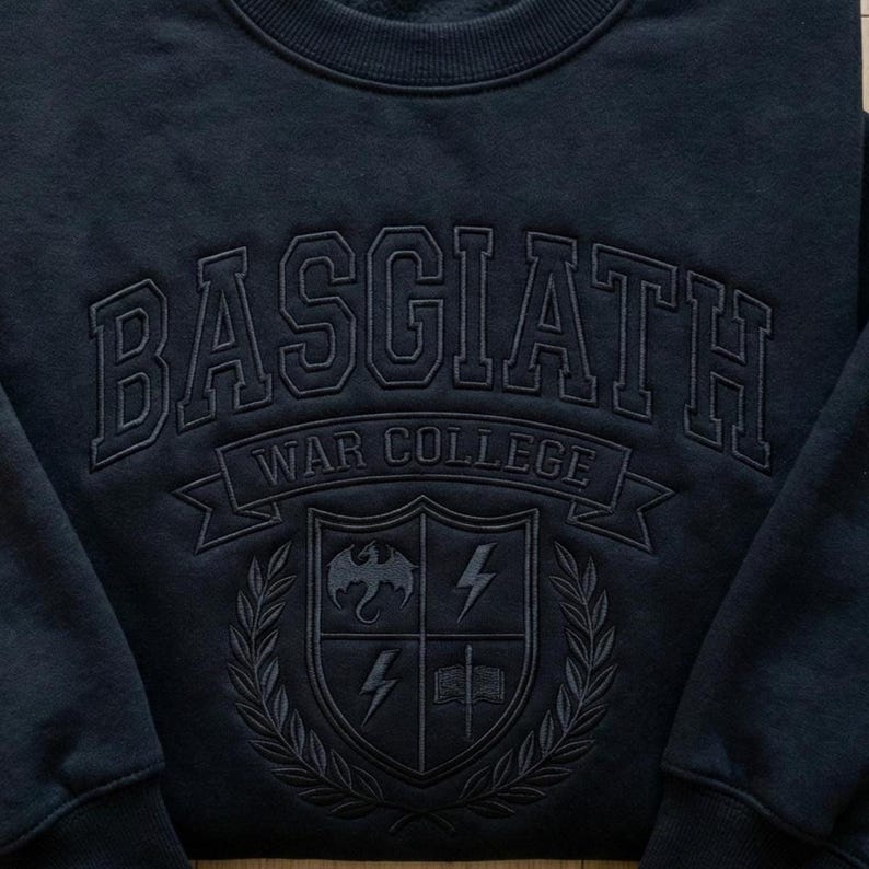 Może przedstawiać: Granatowa bluza z napisem "BASGIATH WAR COLLEGE" w łuku. Poniżej herb z smokiem, piorunami i otwartą książką. Projekt jest w tonacji ton w ton.