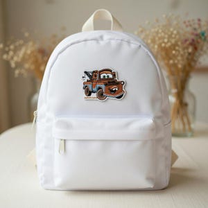 May include: White backpack with a front pocket and a zipper closure. The backpack features a colorful patch of a cartoon tow truck. The patch has the text "custom name" at the bottom.