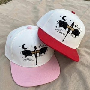 以下が含まれることがあります： ドラゴンのデザインが施された白い野球帽2つ。片方はピンクのつばとボタン、もう片方は赤い色のつばとボタンです。各帽子には、金のアクセントが付いた黒いドラゴン、三日月、星が描かれています。