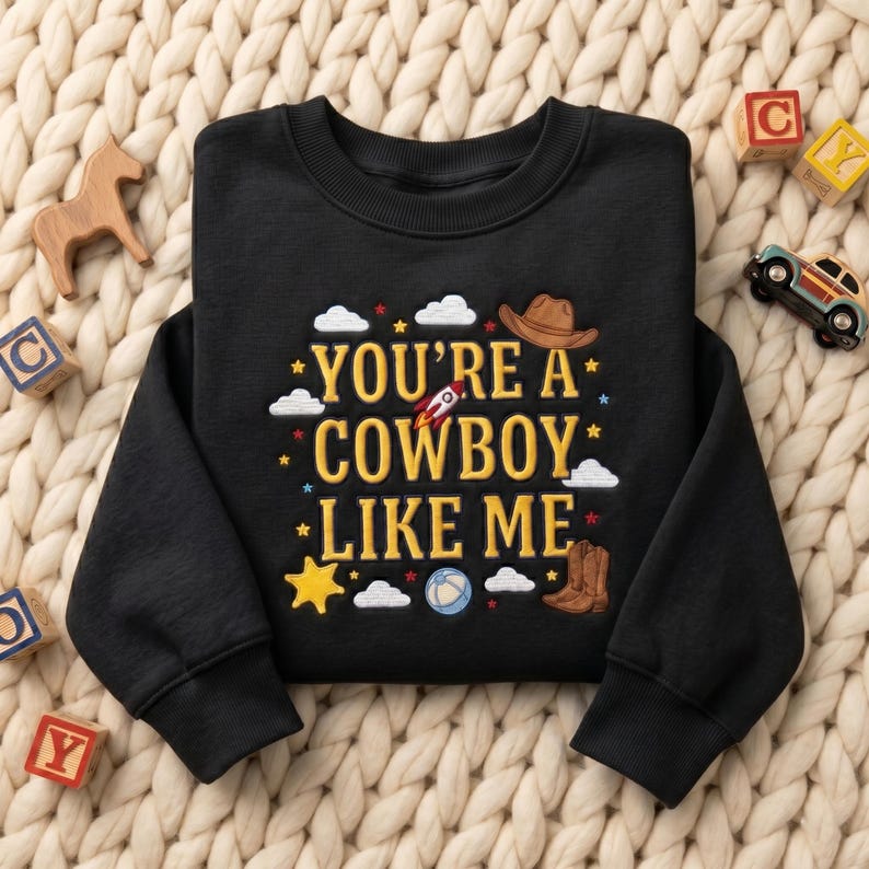 May include: A black long-sleeve sweatshirt featuring yellow embroidered text that reads "YOU'RE A COWBOY LIKE ME." The design includes embroidered motifs of a brown cowboy hat, a rocket ship, a blue ball, and brown cowboy boots, surrounded by white clouds and yellow stars.