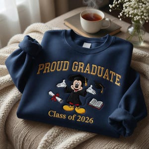 May include: A navy blue sweatshirt with gold embroidered text reading "Proud Graduate" and "Class of 2026". The design features Mickey Mouse in a graduation gown, a diploma, a mortarboard, and an open book.