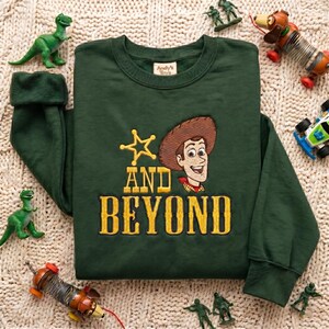 May include: A dark green crewneck sweatshirt featuring an embroidered graphic of a cowboy character wearing a brown hat, a yellow star, and the yellow text "AND BEYOND". The sweatshirt has long sleeves and a ribbed collar, cuffs, and hem.