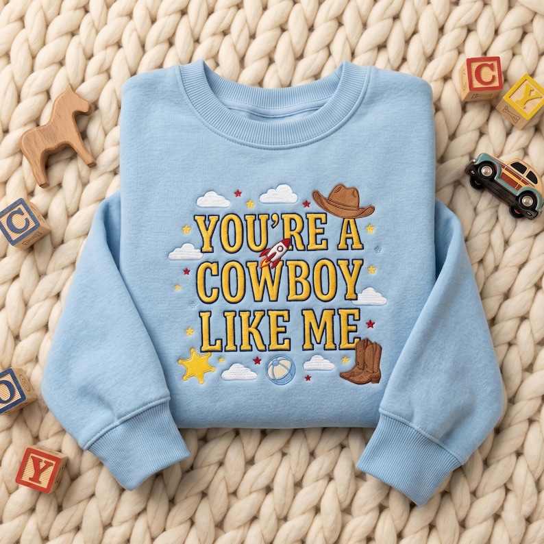 May include: A light blue crewneck sweatshirt featuring yellow embroidered text that reads YOU'RE A COWBOY LIKE ME. The design includes embroidered icons of a brown cowboy hat, a rocket ship, a pair of brown boots, a yellow star, and white clouds.
