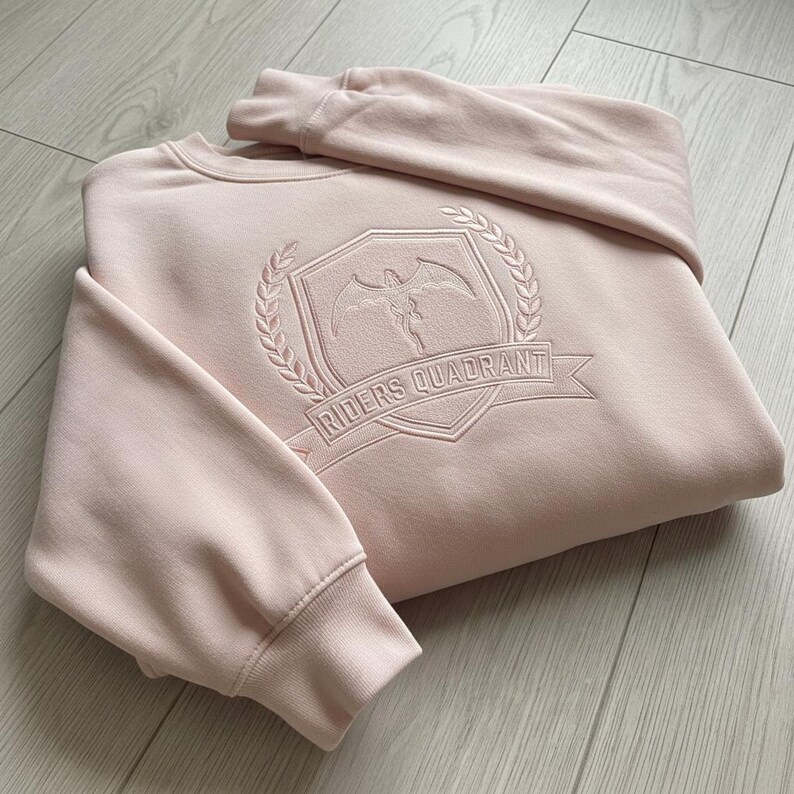 May include: A light pink sweatshirt with the text "RIDERS QUADRANT" embroidered on the front. The design includes a crest with a winged figure and laurel wreath. The sweatshirt has ribbed cuffs and a crew neck.