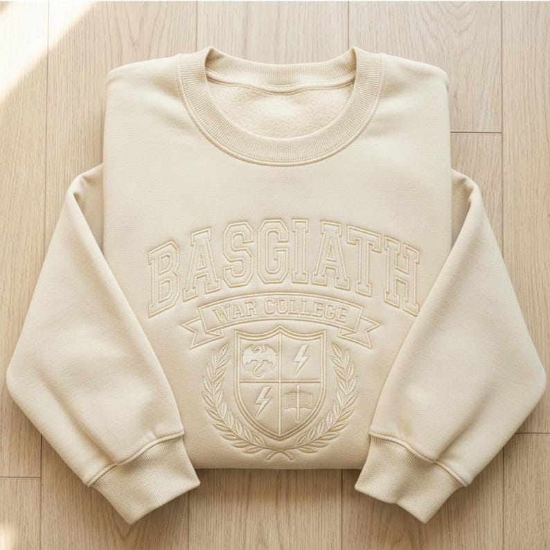 Może przedstawiać: Kremowa bluza z napisem "BASGIATH WAR COLLEGE" w wypukłym, teksturowanym wzorze. Projekt zawiera herb ze smokiem, błyskawicami i banerem. Bluza ma okrągły dekolt i prążkowane mankiety.