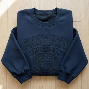 Może przedstawiać: Granatowa bluza z wytłoczonym napisem "BASGIATH WAR COLLEGE" na piersi. Poniżej znajduje się herb z smokiem, błyskawicami i wieńcem laurowym. Bluza jest starannie złożona na drewnianej powierzchni.