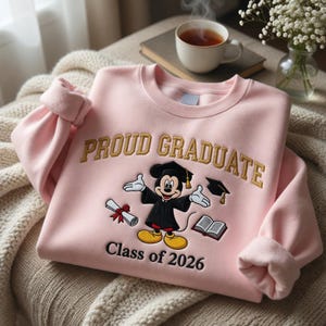 May include: A pale pink sweatshirt embroidered with "PROUD GRADUATE" in gold. The design includes a cartoon Mickey Mouse in a graduation gown, a diploma, a mortarboard, and the text "Class of 2026".