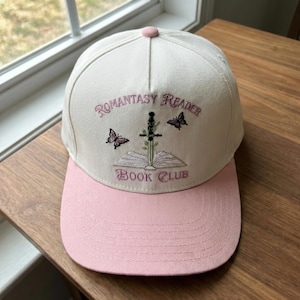 May include: A cream and pink baseball cap with the embroidered text "ROMANTASY READER BOOK CLUB". The cap has a pink peak and button, with butterfly and sword designs. Ideal for book lovers.