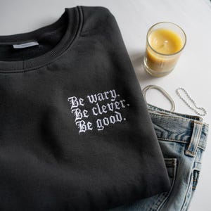 Sudadera bordada "Sé cauteloso, sé inteligente, sé bueno" con estética de versos ensombrecidos e inspiración de fantasía oscura. Cuello redondo con estética literaria.