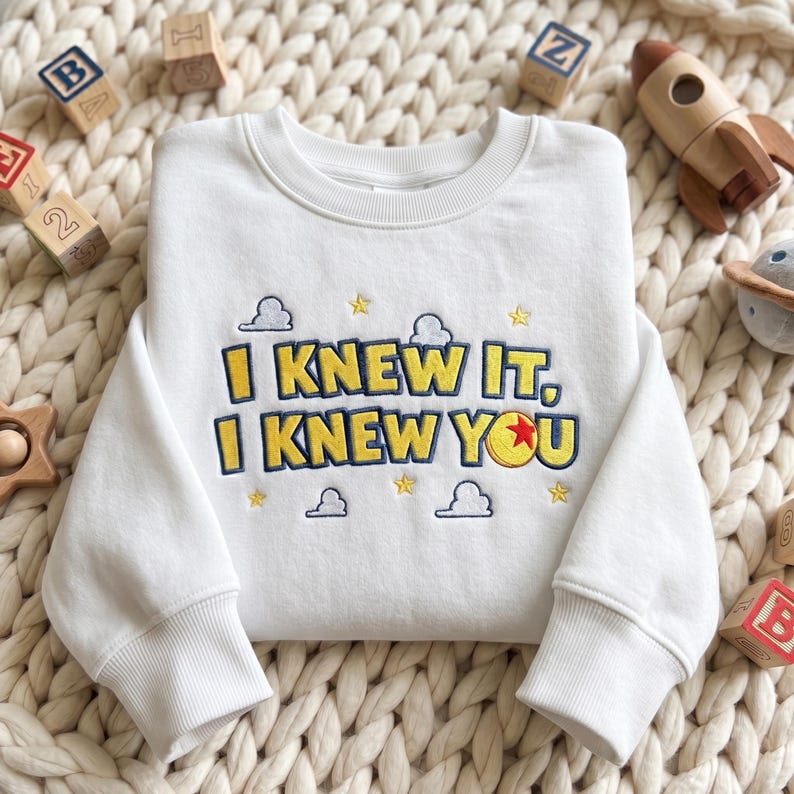 May include: A white crewneck sweatshirt featuring yellow embroidered text that reads I KNEW IT, I KNEW YOU with a blue outline. The design includes small embroidered blue clouds, yellow stars, and a red, yellow, and blue ball icon.