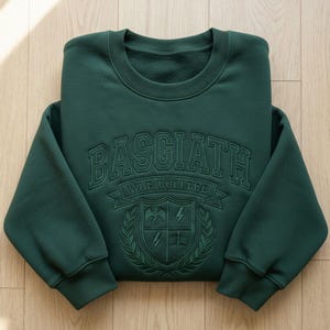 Może przedstawiać: Ciemnozielona bluza z haftowanym napisem "BASGIATH WAR COLLEGE" na piersi. Poniżej herb z smokiem, błyskawicami i książką. Bluza jest złożona na drewnianej powierzchni.
