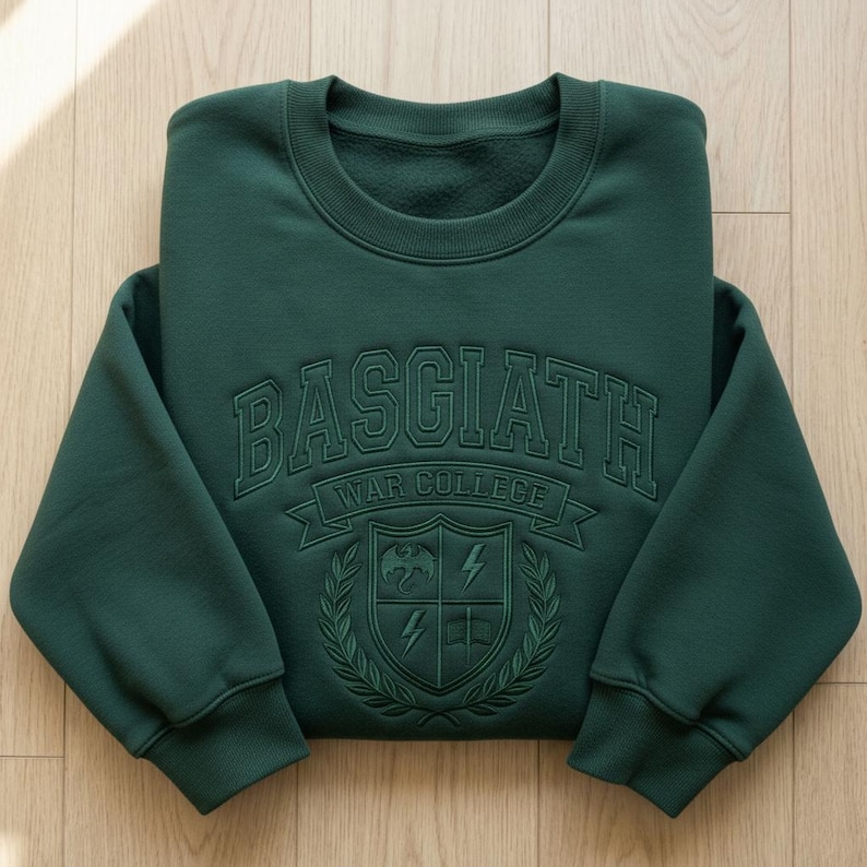 Może przedstawiać: Ciemnozielona bluza z haftowanym napisem "BASGIATH WAR COLLEGE" na piersi. Poniżej herb z smokiem, błyskawicami i książką. Bluza jest złożona na drewnianej powierzchni.