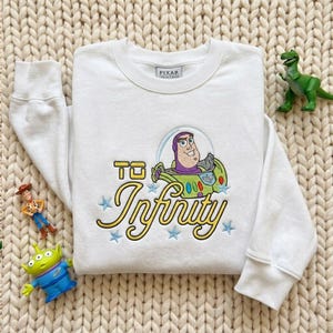 Puede incluir: Una sudadera blanca de manga larga con un bordado de Buzz Lightyear de Toy Story dentro de una burbuja, el texto "TO Infinity" en letra amarilla y peque&ntilde;as estrellas azules. Tiene una etiqueta de Pixar Collectibles en el cuello.
