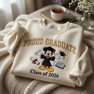 May include: A cream-coloured sweatshirt with gold embroidered text reading "PROUD GRADUATE". It features a Mickey Mouse design in a graduation gown, a diploma, a mortarboard, and a book. The text "Class of 2026" is below the graphic.
