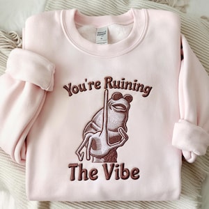 Puede incluir: Sudadera rosa claro con la frase bordada "You're Ruining The Vibe" y una ilustración de rana marrón. La sudadera tiene cuello redondo y mangas largas con puños enrollados.