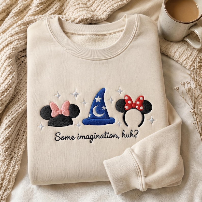 May include: A cream-colored crewneck sweatshirt featuring embroidered designs of black mouse ears with a pink bow, a blue wizard hat with a moon and stars, and black mouse ears with a red polka-dot bow, with the text "Some imagination, huh?"