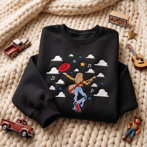 May include: A black crewneck sweatshirt featuring an embroidered design of a person with blonde hair wearing a yellow plaid shirt and blue jeans, holding a guitar and a red hat, surrounded by white clouds, musical notes, and colorful stars.