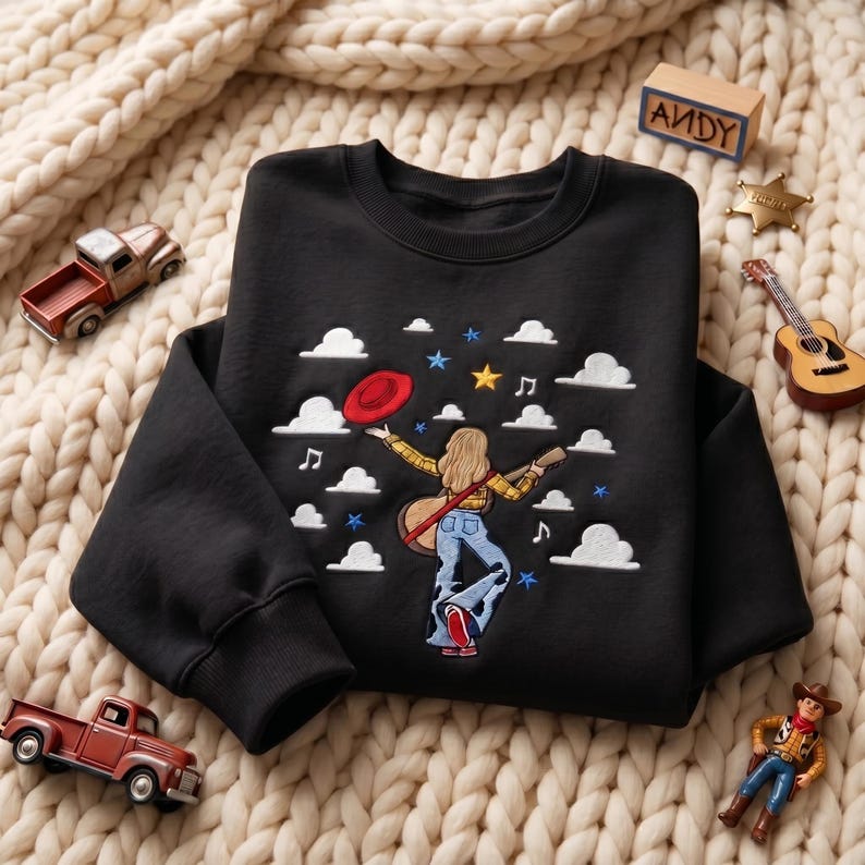 May include: A black crewneck sweatshirt featuring an embroidered design of a person with blonde hair wearing a yellow plaid shirt and blue jeans, holding a guitar and a red hat, surrounded by white clouds, musical notes, and colorful stars.