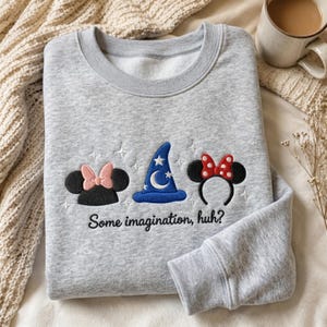 May include: A light gray crewneck sweatshirt featuring embroidered designs of black mouse ears with a pink bow, a blue wizard hat with a white moon and stars, and black mouse ears with a red polka dot bow, with the text "Some imagination, huh?"