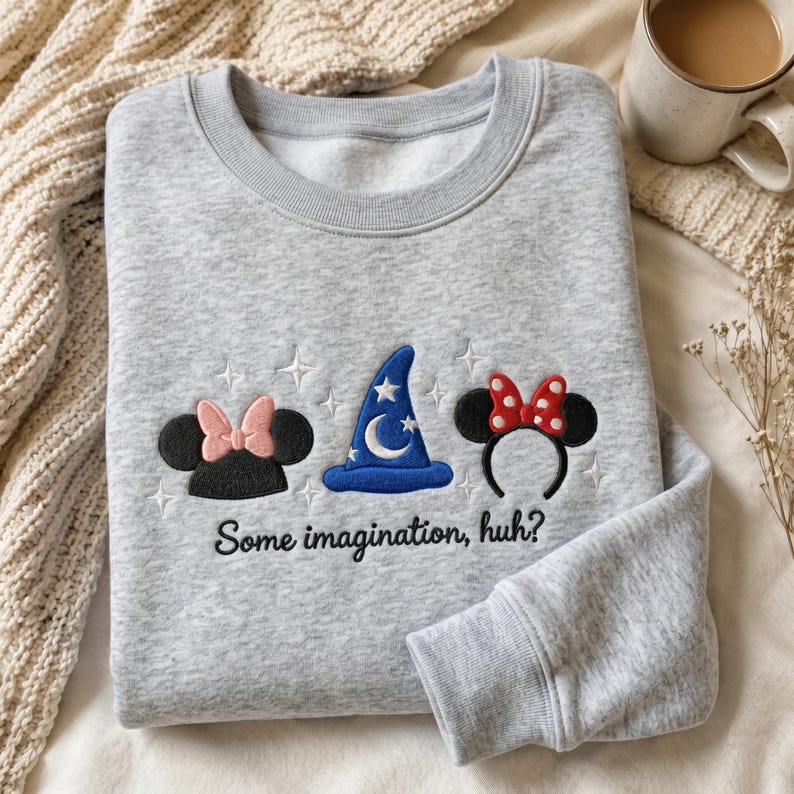 May include: A light gray crewneck sweatshirt featuring embroidered designs of black mouse ears with a pink bow, a blue wizard hat with a white moon and stars, and black mouse ears with a red polka dot bow, with the text "Some imagination, huh?"
