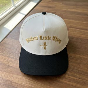 以下が含まれることがあります： 「Violent Little Thing」の文字が金色で刺繍された白と黒の野球帽。つばは黒で、上部に小さな黒いボタンが付いています。テキストの下には金のエンブレムが刺繍されています。