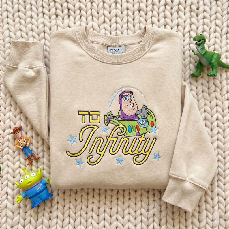 Puede incluir: Una sudadera beige de cuello redondo con un bordado de Buzz Lightyear de Toy Story dentro de una burbuja, junto con el texto amarillo bordado "To Infinity" y varias estrellas peque&ntilde;as bordadas en azul claro alrededor del personaje.