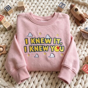 May include: A light pink crewneck sweatshirt featuring embroidered text that reads I KNEW IT, I KNEW YOU in yellow with dark blue outlines. The design includes small embroidered yellow stars and white clouds, with a red and yellow ball icon.
