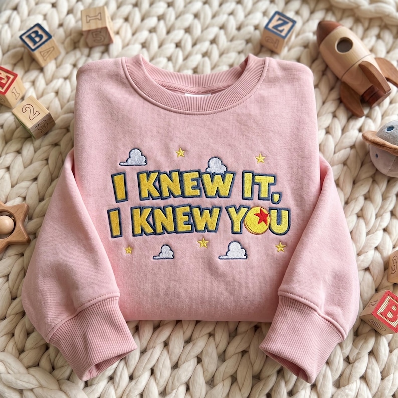 May include: A light pink crewneck sweatshirt featuring embroidered text that reads I KNEW IT, I KNEW YOU in yellow with dark blue outlines. The design includes small embroidered yellow stars and white clouds, with a red and yellow ball icon.