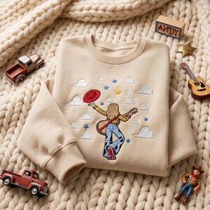 May include: A beige crewneck sweatshirt featuring an embroidered design of a person with long hair wearing a yellow plaid shirt and cow-print pants, holding a guitar and a red hat, surrounded by white clouds, blue stars, and musical notes.