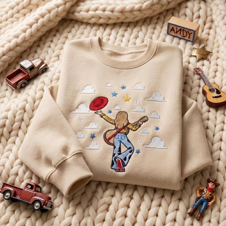 May include: A beige crewneck sweatshirt featuring an embroidered design of a person with long hair wearing a yellow plaid shirt and cow-print pants, holding a guitar and a red hat, surrounded by white clouds, blue stars, and musical notes.