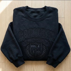Może przedstawiać: Granatowa bluza z napisem "BASGIATH WAR COLLEGE" w wypukłym, teksturowanym wzorze. Poniżej herb z smokiem, piorunami i wieńcem laurowym. Bluza jest złożona na drewnianej powierzchni.