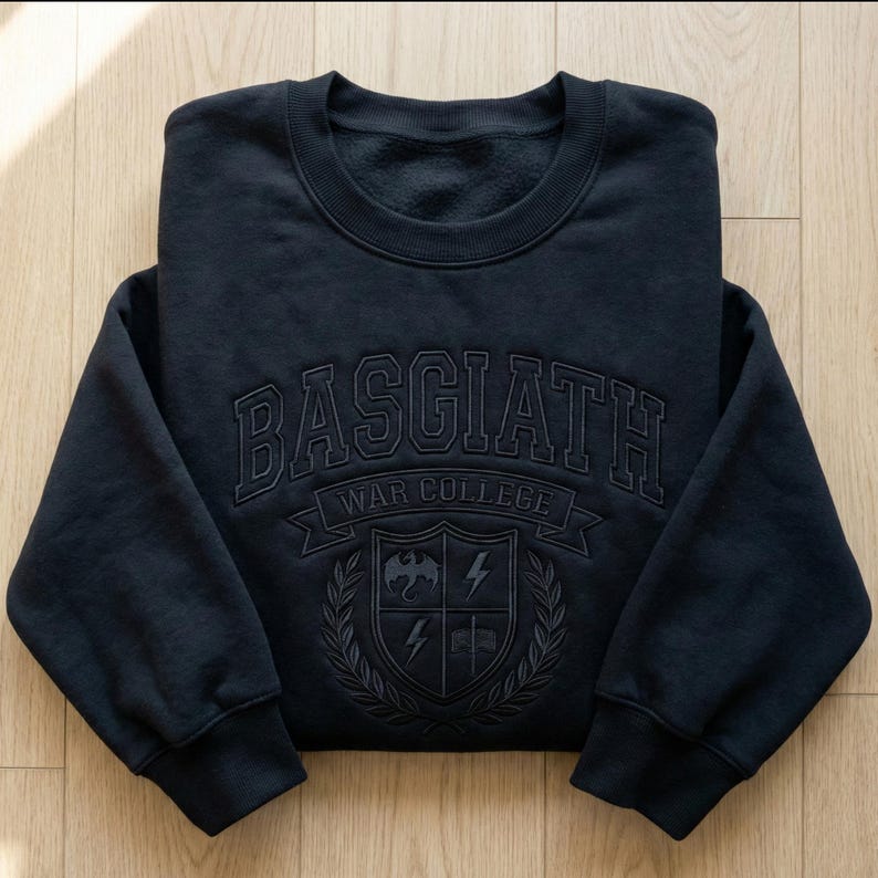Może przedstawiać: Granatowa bluza z napisem "BASGIATH WAR COLLEGE" w wypukłym, teksturowanym wzorze. Poniżej herb z smokiem, piorunami i wieńcem laurowym. Bluza jest złożona na drewnianej powierzchni.