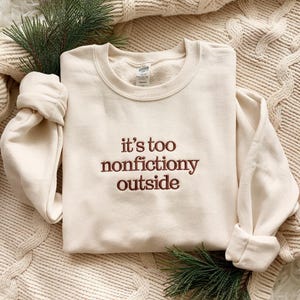 May include: Cream-colored sweatshirt with the embroidered phrase "it's too nonfictiony outside" in a brown color. The long-sleeved top is folded on a textured surface, with a sprig of greenery.
