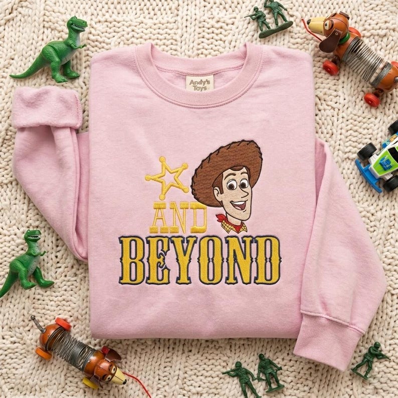 May include: A light pink crewneck sweatshirt featuring an embroidered graphic of a cowboy character with a brown hat and red neckerchief, a yellow star, and the yellow text "AND BEYOND" in a bold, western-style font on the front chest.