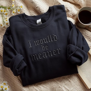 May include: A dark navy sweatshirt featuring the embroidered phrase "I would be meaner." The top is displayed on a cream-colored blanket, with a cup of coffee and a book in the background.