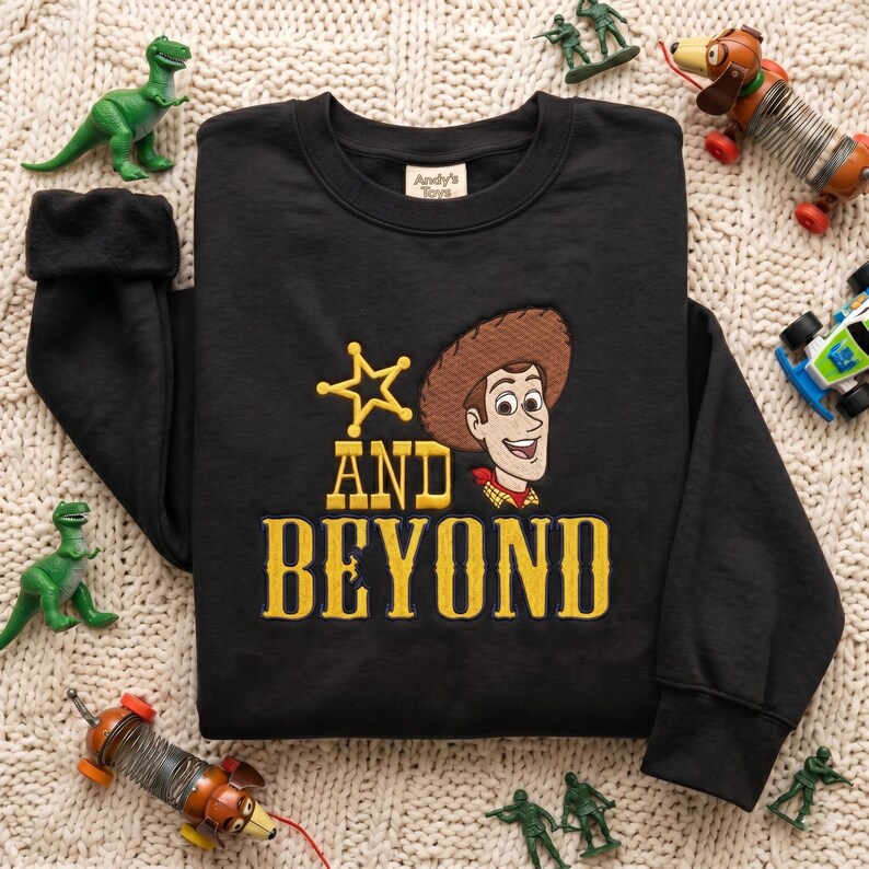 May include: A black long-sleeve sweatshirt featuring an embroidered graphic of a cowboy character wearing a brown hat and red neckerchief, a yellow star, and the yellow text "AND BEYOND". The inner neck label reads "Andy's Toys".
