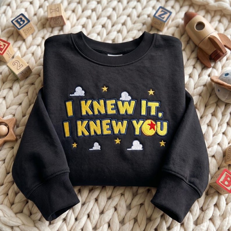 May include: A black long-sleeve sweatshirt featuring yellow embroidered text that reads I KNEW IT, I KNEW YOU. The design includes small white clouds and yellow stars, with a red, yellow, and blue ball replacing the letter O in YOU.