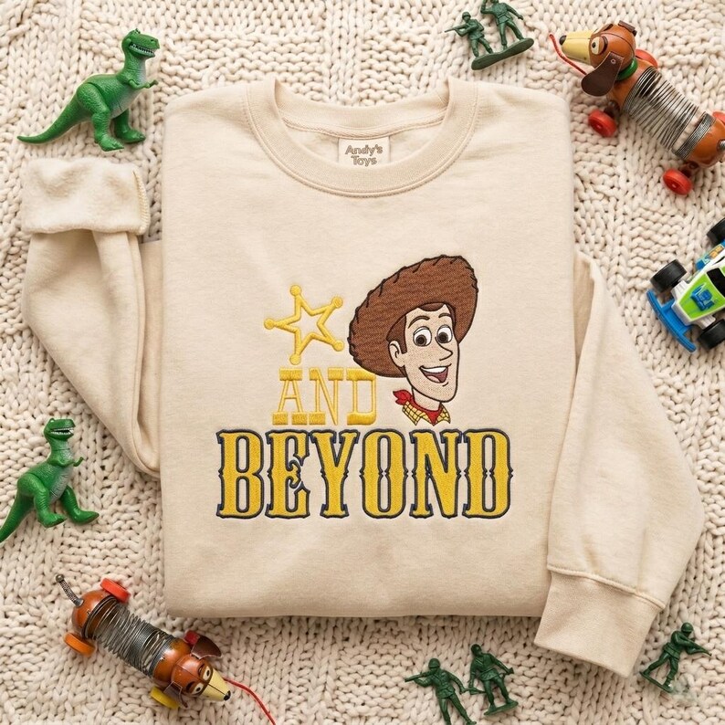 May include: A beige crewneck sweatshirt featuring an embroidered graphic of a cowboy character wearing a brown hat, a yellow star, and the yellow text "AND BEYOND" in a western-style font. The interior neck label reads "Andy's Toys".