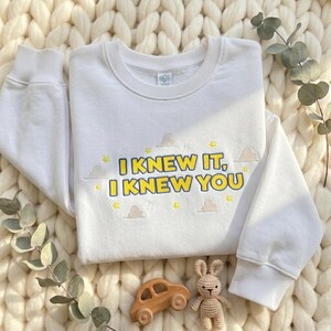 May include: A white long-sleeve sweatshirt featuring the embroidered text "I KNEW IT, I KNEW YOU" in yellow with a dark blue outline. The design includes small embroidered yellow stars, light brown clouds, and musical notes surrounding the text.