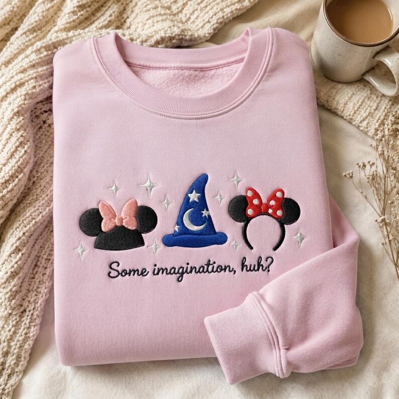 May include: A light pink crewneck sweatshirt featuring embroidered designs of black mouse ears with a pink bow, a blue wizard hat with a white moon and stars, and black mouse ears with a red polka dot bow, with the text Some imagination, huh?