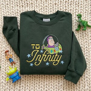 Puede incluir: Una sudadera de manga larga de color verde oscuro con un bordado de Buzz Lightyear dentro de un casco de burbuja. El texto "TO Infinity" aparece en letras amarillas, decorado con estrellas bordadas en azul claro.