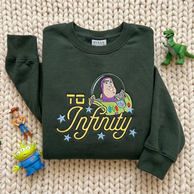 Puede incluir: Una sudadera de manga larga de color verde oscuro con un bordado de Buzz Lightyear dentro de un casco de burbuja. El texto "TO Infinity" aparece en letras amarillas, decorado con estrellas bordadas en azul claro.