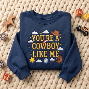 May include: A navy blue long-sleeve sweatshirt featuring yellow embroidered text that reads "YOU'RE A COWBOY LIKE ME." The design includes embroidered motifs of a brown cowboy hat, a pair of brown boots, a rocket ship, clouds, stars, and a ball.