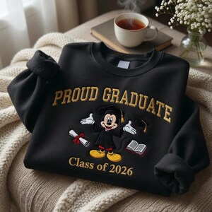 May include: Black sweatshirt with gold embroidered text reading "PROUD GRADUATE" and "Class of 2026". Features a cartoon Mickey Mouse in a graduation gown, holding a diploma and a graduation cap. A book is also shown.