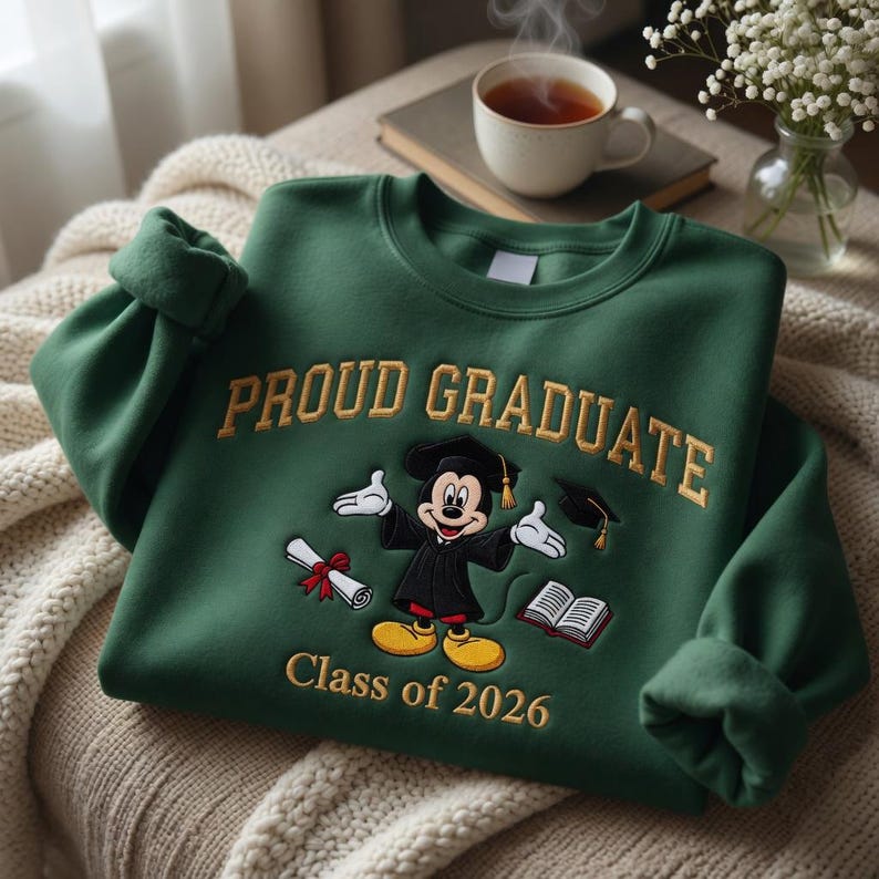 May include: A dark green sweatshirt featuring gold embroidered text reading "PROUD GRADUATE". The design includes a cartoon character in a graduation gown, a diploma, and "Class of 2026".