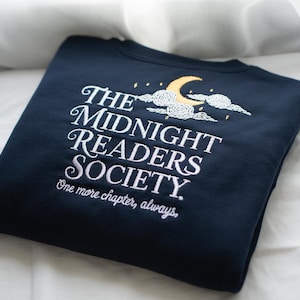 Puede incluir: Sudadera azul marino con texto bordado en blanco: "The Midnight Readers Society. One more chapter, always." Presenta un diseño de luna creciente y nubes. Una prenda cómoda para amantes de la lectura.