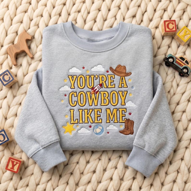 May include: A light gray crewneck sweatshirt featuring yellow embroidered text that reads YOU'RE A COWBOY LIKE ME. The design includes embroidered icons of a brown cowboy hat, a rocket ship, a yellow star, a blue ball, and brown cowboy boots.