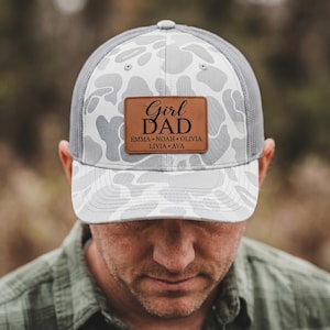 Puede incluir: Gorra de camionero gris y blanca con estampado de camuflaje y parche de cuero marrón. El parche dice "Girl Dad" con los nombres Emma, Noah, Olivia, Livia y Ava. La gorra tiene una parte trasera de malla gris.