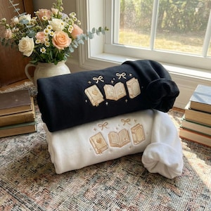 Puede incluir: Dos sudaderas, una negra y otra blanca, están apiladas. Cada una tiene un diseño bordado de libros, corazones y lazos. Las sudaderas están dobladas sobre una alfombra estampada, con un jarrón de flores en el fondo.