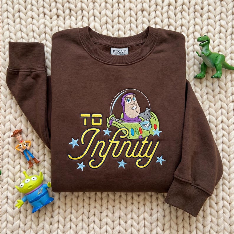 Puede incluir: Una sudadera de cuello redondo en color marr&oacute;n oscuro con un bordado de Buzz Lightyear de Toy Story. El texto "TO Infinity" aparece en letras amarillas, decorado con estrellas bordadas en azul claro y una etiqueta de Pixar.